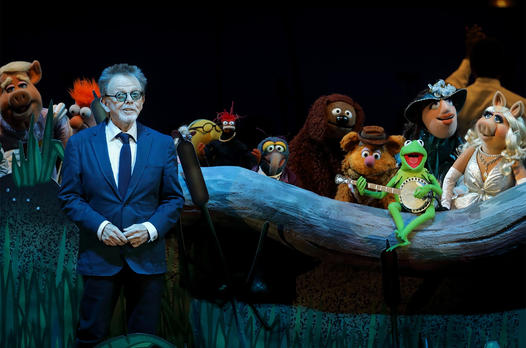 A man in a suit standing in front of a group of muppets.