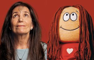 OH – Celebrating 50 Years of Cathy | Aug 8th A woman with long brown hair looks up next to a smiling doll with long red yarn hair, both against a red background.