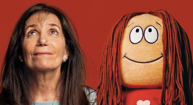 OH – Celebrating 50 Years of Cathy | Aug 8th A woman with long brown hair looks up next to a smiling doll with long red yarn hair, both against a red background.