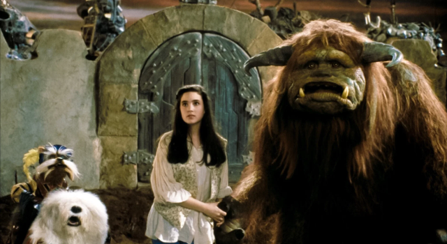 SF – Celebrate 40 Years of Labyrinth | Aug 22nd A young woman stands next to a large horned creature and two fantasy animals in front of a stone door in a fantasy setting.