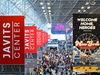 NYCC 20th Anniversary Event Draws +250K Attendees A crowded Javits Center atrium during New York Comic Con, with red vertical banners and a large event sign hanging overhead.