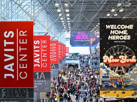 NYCC 20th Anniversary Event Draws +250K Attendees A crowded Javits Center atrium during New York Comic Con, with red vertical banners and a large event sign hanging overhead.